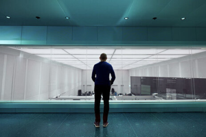 A silhouette of a man looking down at a clean, white office space through glass windows.