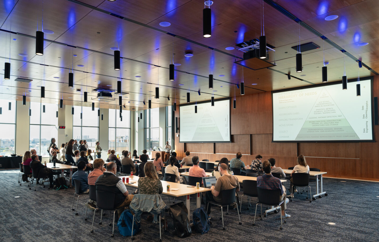 A group of people in a lecture room listening to a speaker. There are blue lights overhead and large, bright windows in the background.
