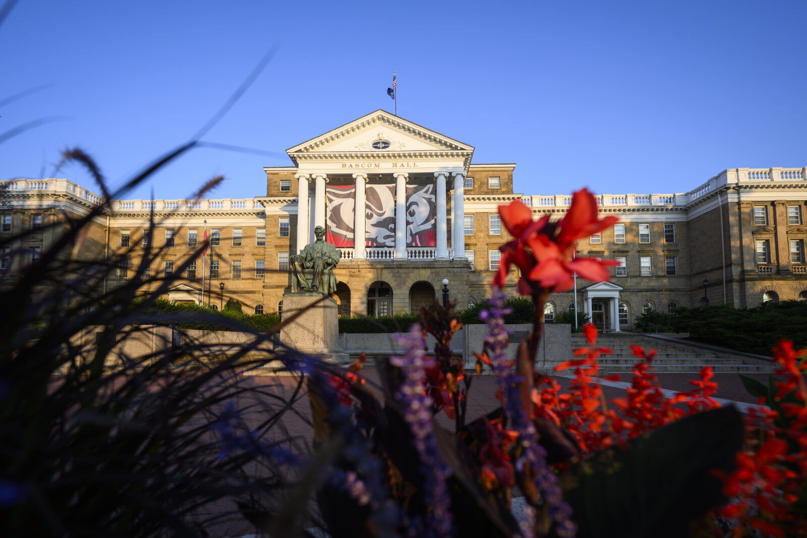 Picture of Bascom Hall featuring blue sky. The building is semi-covered by grass along with red and purple flowers.