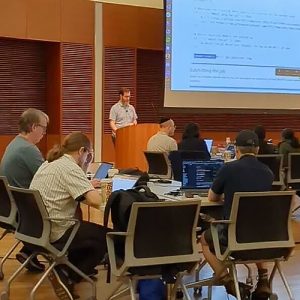 An instructor standing in front of a class. There is a projection on a screen behind him.