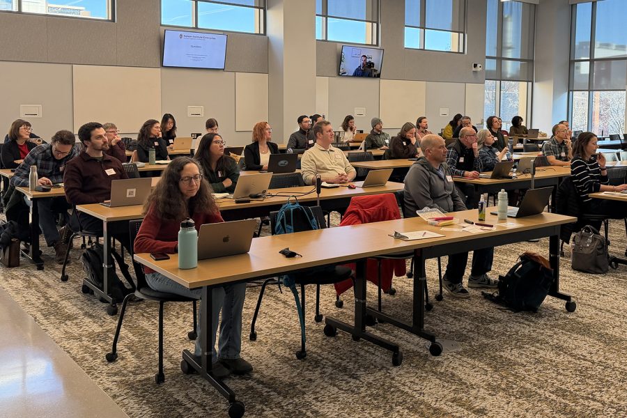 People sitting in a classroom. The tables are arranged in rows. The people are listening to a speaker.